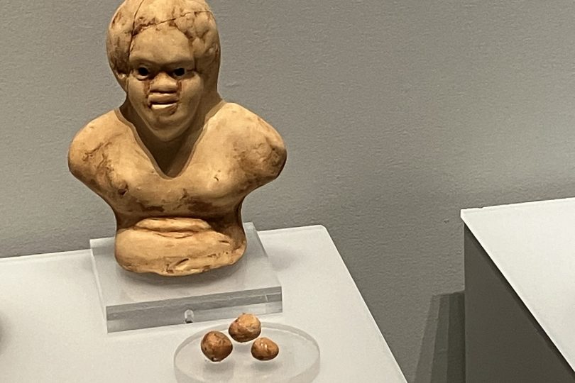 A child's rattle in the shape of a human torso and head. It is made from baked clay and has three of its beads next to it in its museum case.