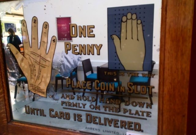 Part of an early twentieth century pier or arcade machine. The sign promises to read the user's palm for one penny, and gives instructions.